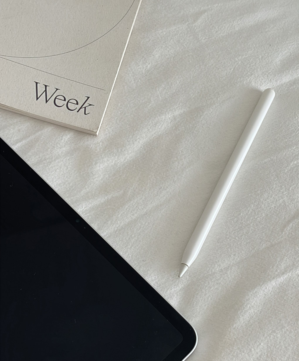 A weekly planner, an iPad, and an Apple Pencil on a white bedsheet